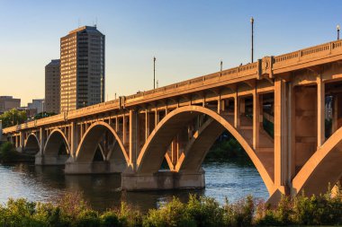 Bir köprü, arka planda bir şehir olan bir nehri kapsar. Köprü betondan yapılmıştır ve büyük bir kemeri vardır.