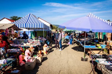 Mavi ve beyaz çizgili çadırı ve mavi şemsiyesi olan bir pazar. İnsanlar alışverişte ve eşyaları karıştırıyor