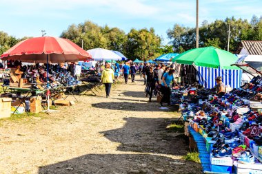 Bir grup insan bir sıra çadırın önünde bir patikada yürüyorlar. Çadırlar, büyük kırmızı bir şemsiye de dahil olmak üzere çeşitli boyutlarda ve renklerdedir. İnsanlar satılık ürünlere göz atıyorlar.