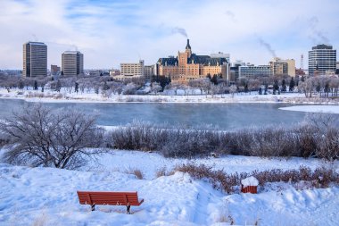 A bench is sitting in the snow next to a river. The bench is red and is located in a park. The park is surrounded by buildings and trees. The snow is covering the ground and the sky is cloudy clipart