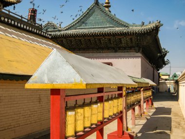 Bir sıra sarı ve kırmızı dua tekerlekleri bir binanın önünde dizilmiştir. Arka planda kuşlar uçuşurken sahne huzurlu ve huzurlu.