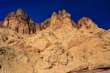 Arka planda mavi gökyüzü olan bir dağ sırası. Dağlar kahverengi ve kayalıktır. Gökyüzü açık ve parlak
