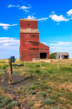 Bir tarlanın ortasında kırmızı tahıl silosu var. Silo bir çitle çevrilidir ve bölgedeki tek binadır. Gökyüzü açık ve mavidir ve güneş ışıl ışıl parlar.