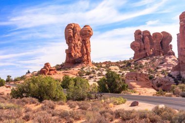 Bir yolu ve birkaç büyük kayası olan bir çöl manzarası. Gökyüzü mavi ve bazı bulutlar var.