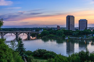 Üzerinden nehir akan bir şehrin üzerinde güzel bir gün batımı. Gökyüzü bulutlarla dolu ve su sakin. Arka plandaki binalar yüksek ve köprü görünür.