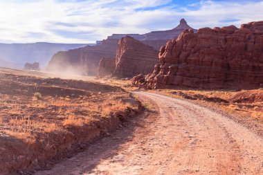 Arka planda bir dağ olan toprak bir yol çöl manzarası boyunca esiyor. Yol tozlu ve gökyüzü bulutlu.