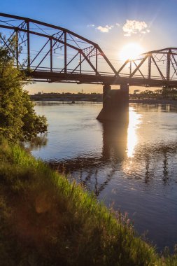 Bir köprü, suyun üzerinde parlayan bir güneş ile bir nehri kaplar. Köprü eski ve kırsal bir görünüme sahip.
