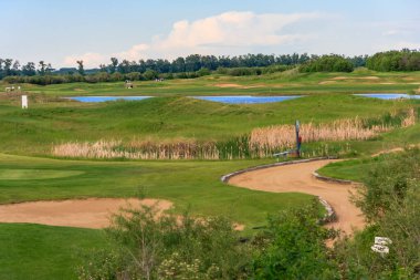 Arka planda gölet olan bir golf sahası. Gölet çimenler ve ağaçlarla çevrilidir. Golf sahası iyi korunuyor ve göl manzarası da çok güzel.