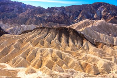 Arkasında dağ sıraları olan bir çöl manzarası. Tepeler toprak ve kayalarla kaplı. Bu da sahneyi engebeli ve çorak bir hale getiriyor. Gökyüzü açık ve mavi