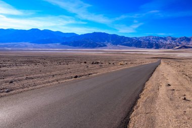 Arka planda dağlar olan çölde bir yol. Gökyüzü mavi ve güneş parlıyor.