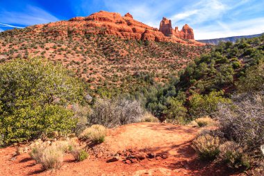 Kayalık bir tepesi ve toprak yolu olan bir çöl manzarası. Gökyüzü açık ve güneş parlıyor.