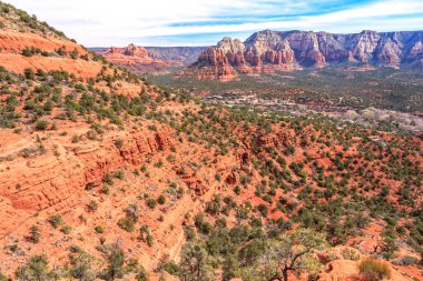 Arkasında dağ sıraları olan bir çöl manzarası. Dağlar kırmızı kayalarla kaplı ve çöl ağaçlarla dolu. Gökyüzü açık ve güneş parlıyor.