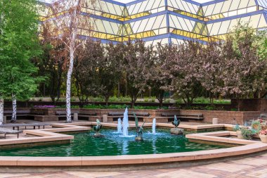 Arka planda büyük bir bina olan bir çeşme. Çeşme banklar ve ağaçlarla çevrilidir. Manzara huzurlu ve rahatlatıcı.