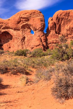 Arkasında büyük bir kaya oluşumu olan bir çöl manzarası. Gökyüzü mavi ve güneş parlıyor.