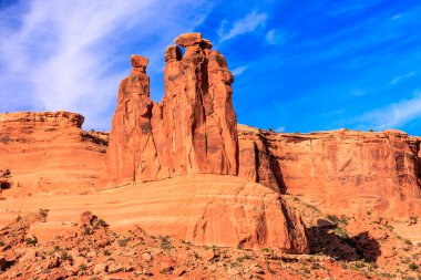Üzerinde iki küçük kaya olan büyük bir kaya oluşumu. Kayalar kayalık bir yamaçla çevrilidir ve gökyüzü açık ve mavidir. Sahnenin ihtişam ve ihtişam duygusu var.