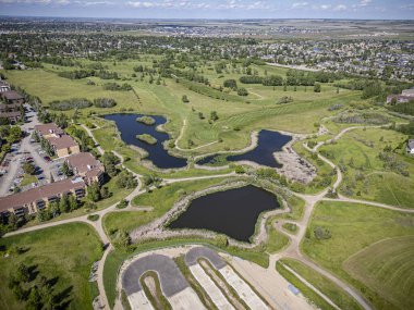 Arka planda birkaç ev ve göleti olan bir golf sahası manzarası. Gölet çimenler ve ağaçlarla çevrili ve golf sahası kırsal bir alanda yer alıyor.