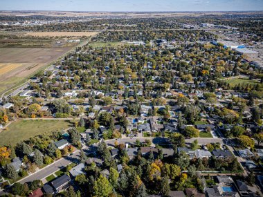 Evleri ve ağaçları olan bir yerleşim yeri manzarası. Evler çoğunlukla kahverengi ve beyazdır ve ağaçlar çoğunlukla yeşildir. Gökyüzü mavi ve hiç bulut yok.