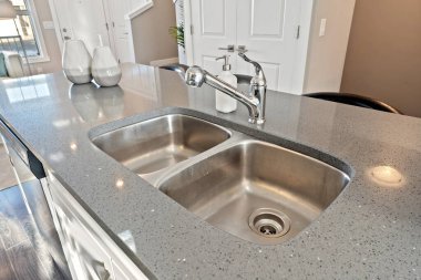 Gümüş musluğu olan paslanmaz çelik bir lavabo tezgahın üzerinde duruyor. Lavabo büyük ve iki bölmesi var. Tezgah granitten yapılmış ve modern bir tasarımı var. Lavabo ışıkla çevrili.