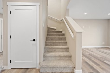 Siyah saplı ve siyah düğmeli beyaz bir kapı. Kapı halı döşeli bir merdivenin hemen yanında. Oda boş ve minimalist bir görünüşü var.