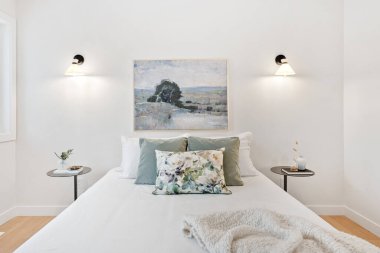 Büyük yataklı ve iki komodinli beyaz bir yatak odası. Yatak beyaz bir yorgan ve yastıkla kaplı ve üzerinde çerçeveli bir tablo asılı. Oda temiz.