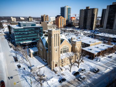 Arkasında büyük bir kilise olan Karlı şehir caddesi. Kilise yüksek binalarla çevrilidir ve sahnenin odak noktasıdır. Karla kaplı zemin ve arabaların varlığı