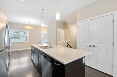 Beyaz bir adası ve paslanmaz çelik aletleri olan bir mutfak. Mutfak temiz ve iyi ışıklandırılmış, geniş bir pencere doğal ışık sağlıyor. Buzdolabı odanın sol köşesinde.