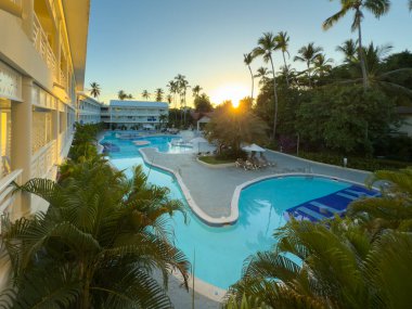 Arka planda günbatımı olan güzel bir havuz alanı. Havuz alanı palmiye ağaçlarıyla çevrili ve rahatlatıcı bir atmosferi var.