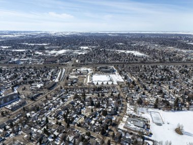 Şehir manzarası, yerde kar ve evler. Evlerin çoğu beyaz ve bazı kahverengi evler de var. Gökyüzü mavi ve bazı bulutlar var.