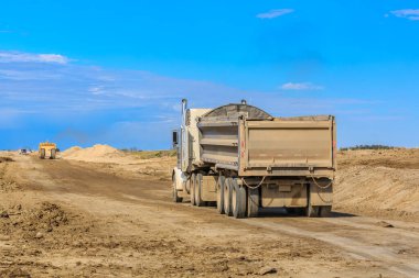 Çöp kamyonu toprak bir yolda ilerliyor. Kamyon bir sürü pislik taşıyor. Gökyüzü mavi ve hiç bulut yok.