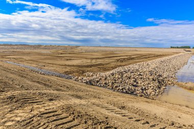 Geniş arazi toprak ve kayalarla kaplıdır. Gökyüzü mavi ve arka planda bulutlar var.