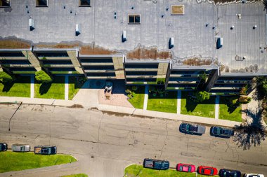 Arka planda binalar olan bir otopark manzarası. Binalar uzun ve modern bir tasarıma sahip. Park yerinde birkaç araba var, biri kırmızı, biri siyah.