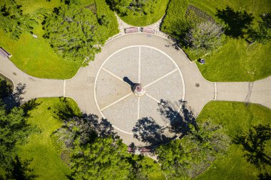Ortasında büyük yuvarlak taştan bir yapı olan park. Taş yapı ağaçlar ve otlarla çevrilidir. Yapının etrafında banklar var ve arka planda birkaç kişi görünüyor.