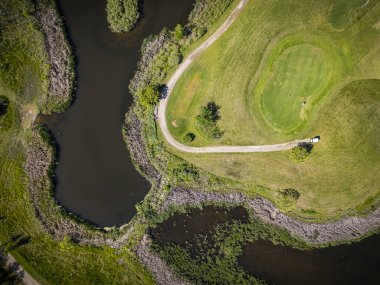 Göleti ve yolu olan golf sahası. Gölet ağaçlar ve otlarla çevrilidir. Göletin yanında bir golf arabası var.