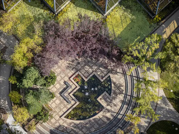 Etrafı bitkilerle çevrili büyük bir çeşme. Bitkiler yeşil ve sarı, çeşme ise mavi. Çeşme bir parkın ortasında ve etrafında ağaçlar var.