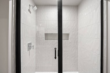 Duşu ve cam kapısı olan bir banyo. Duşun beyaz fayans duvarı ve siyah sapı var.