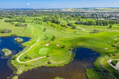 Arka planda gölet olan golf sahası. Gölet çimenler ve ağaçlarla çevrilidir. Golf sahası, uzaktaki evleri olan kırsal bir alanda yer almaktadır.