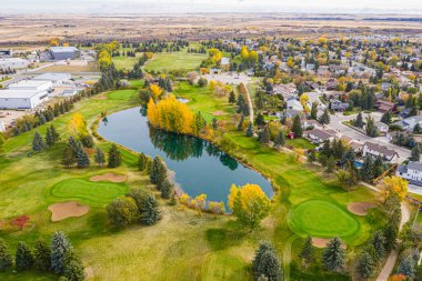 Göllü golf sahası ve arka planda bir yerleşim alanı. Gölet ağaçlarla çevrili ve golf sahasının yeşil çimenli bir alanı var.