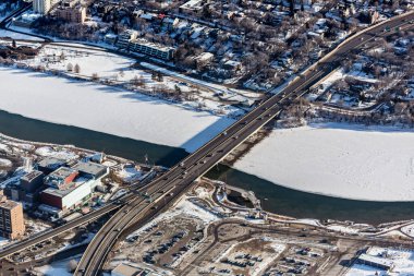 Nehir üzerinde köprüsü olan bir şehir caddesi. Köprü arabalar ve kamyonlarla dolu. Şehir karla kaplı.