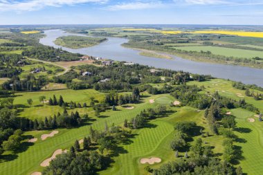 Arka planda nehir olan golf sahası. Golf sahası ağaçlarla çevrili ve yeşil çimenli bir alanı var.