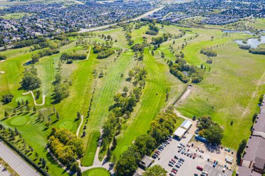Yeşil çimenlikli bir golf sahası ve arabalı bir otopark. Otopark arabalarla dolu ve etrafta dolaşan birkaç kişi var.