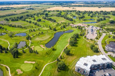 Arka planda büyük bir bina olan golf sahası. Golf sahası ağaçlarla çevrili ve bir göleti var.