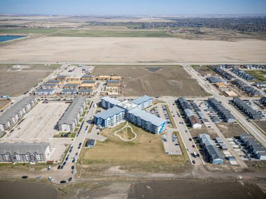 Şehir manzarası ve ortasında büyük bir bina. Binaların çoğu beyaz ve mavi. Otoparka park etmiş bir sürü araba var.