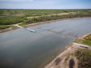 Feribot çimenli bir bölgenin yakınındaki bir nehirde yüzüyor. Gökyüzü açık ve su sakin.