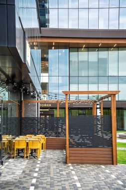 Ahşap çitleri ve sarı sandalyeleri olan restoran avlusu. Veranda, cam cepheli büyük bir binanın önünde yer almaktadır.