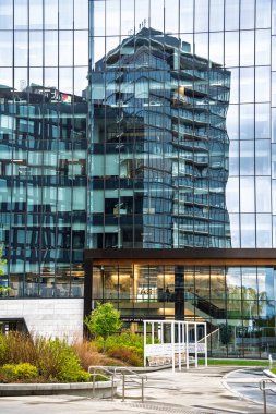 Önünde cam bir cephe ve yeşil bir alan olan yüksek bir bina. Bina cam bir geçitle çevrili.