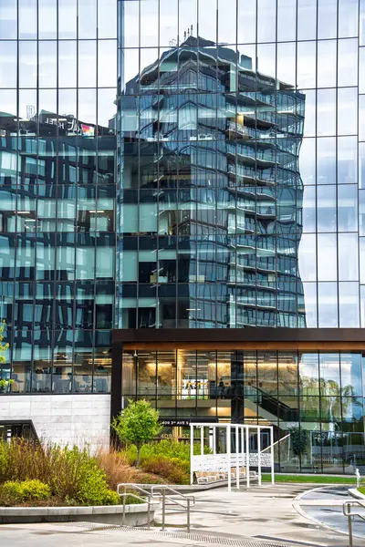 Önünde cam bir cephe ve yeşil bir alan olan yüksek bir bina. Bina cam bir geçitle çevrili.