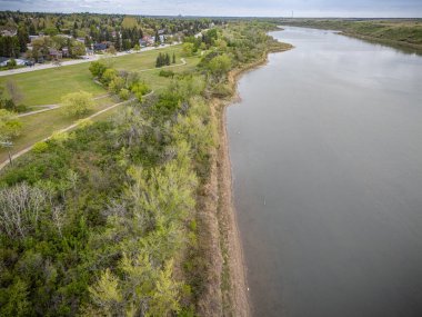 Nehir, arka planda bir kasaba olan ormanlık bir bölgeden geçiyor. Su sakin, ağaçlar yemyeşil.
