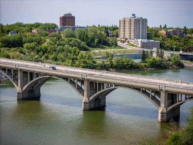 Köprü, arka planda bir şehir bulunan bir nehri kapsar. Köprü yeşil renkli, büyük, gri bir yapıdır. Su sakin ve gökyüzü berrak.