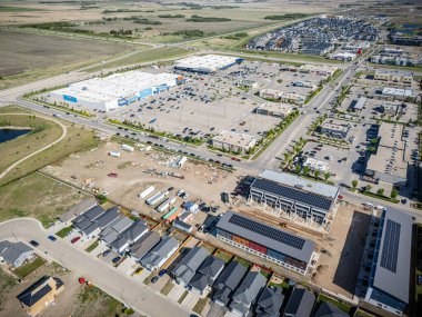 Arka planda büyük bir alışveriş merkezi olan bir yerleşim yeri manzarası. Alışveriş merkezi araba ve kamyonlarla dolu ve bölgede birkaç bina var. Sahne meşgul ve hareketli.