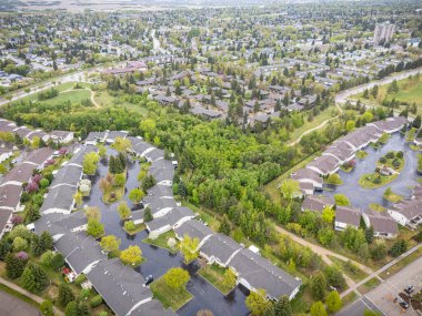 Evleri ve ağaçları olan bir yerleşim alanı. Evlerin çoğu beyaz ve caddelerde park edilmiş birçok araba var. Gökyüzü bulutlu ve görüntünün genel havası sakin ve huzurlu.